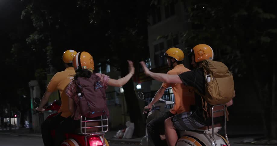 Four people ride on two motorcycles and the two people in the back high five
