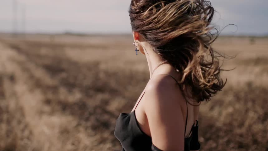 Woman in a black dress stands in a field as the wind blows her hair