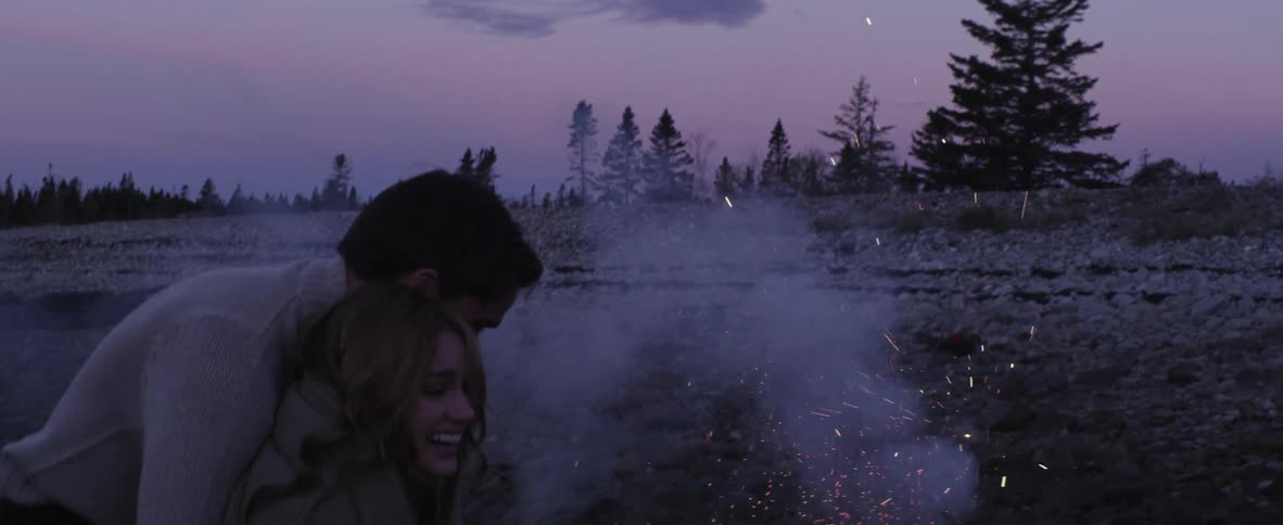 A man and a woman stand in a field cuddling and laughing as sparks fly up from the ground