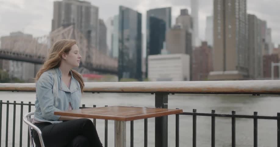 Woman sits at a table next to a river and skyscrapers