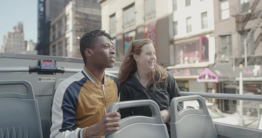 Man and woman sit on top of a tour bus in the city on a sunny day