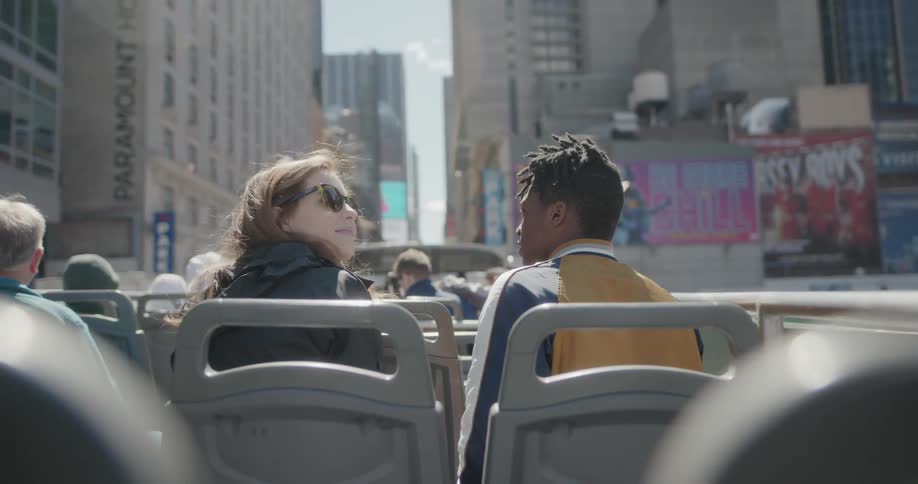 Man and woman sit on top of a tour bus in the city on a sunny day