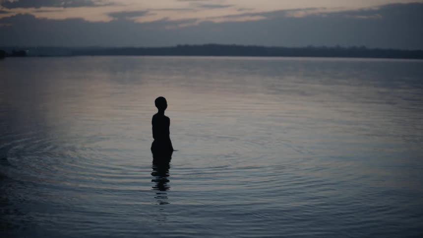 A boy stands in a rippling lake at dusk