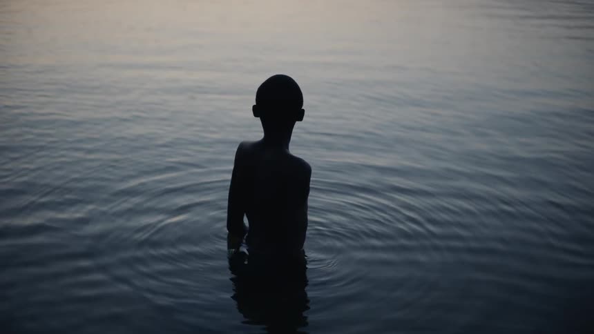 A boy is standing waste-deep in a lake at dawn