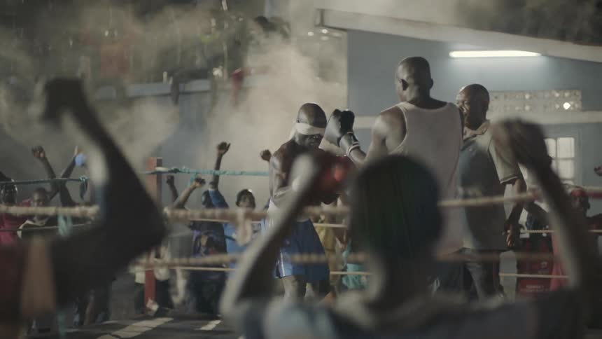 Man wears a blindfold in a boxing ring and people around cheer and wave their arms