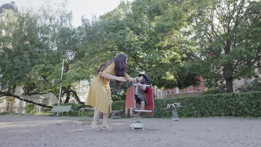 A mother bending down to assist her child on a playground rocker.
