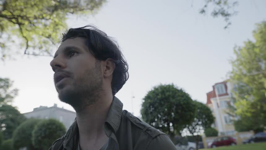 A young man looking away and talking while standing outdoors in a park.