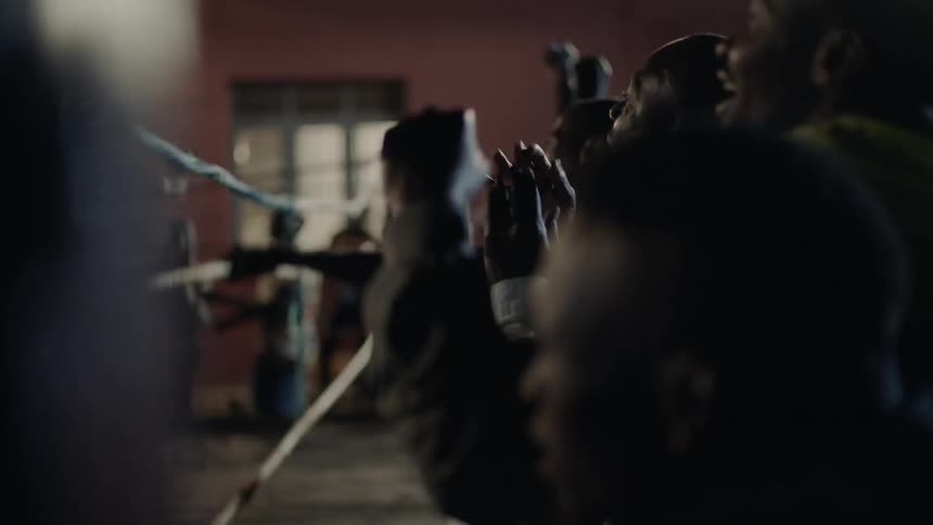 Audience of men cheer and shake the ropes of a boxing ring