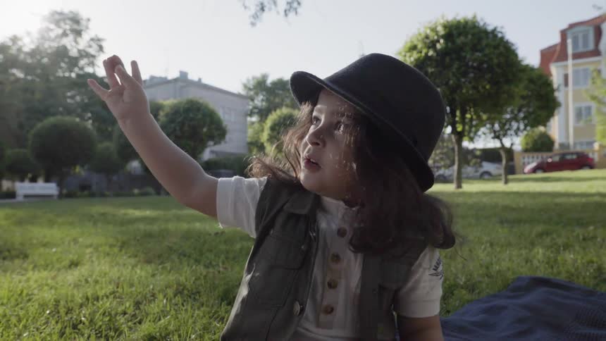A cute child wearing a hat reaching up while sitting outdoors in park.