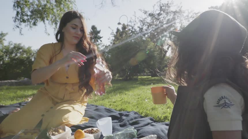 A mother pouring juice into a cup held by her young child outdoors.