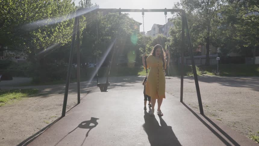 A young child pushing mother on a swing set at park playground.