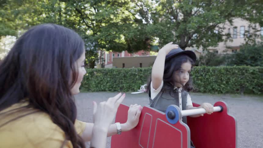 A mother waving playfully to her child riding on a spring rocker playground.