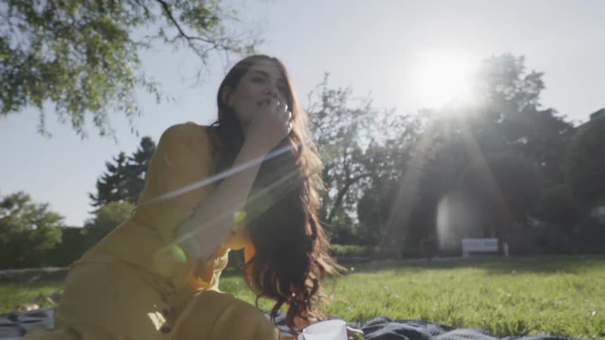 A young woman laughing while sitting outdoors on grass during a sunny picnic.