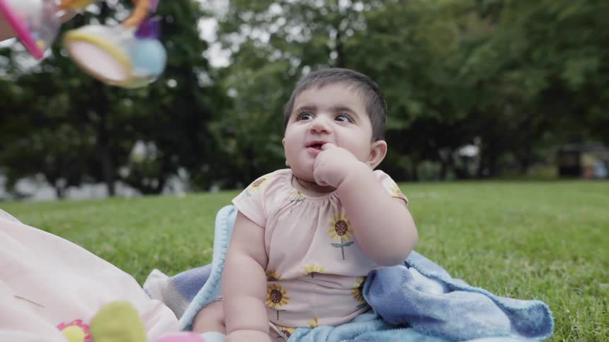 Cute joyful baby sucking thumb while sitting on a picnic blanket outdoors.