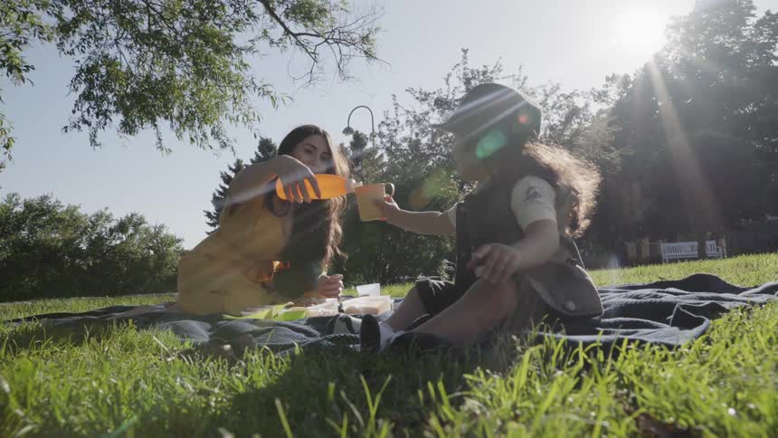A mother pouring orange juice into cup held by child at picnic.