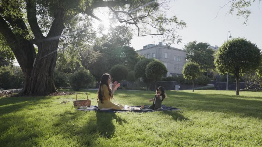 A mother and child sitting on blanket clapping hands during park picnic.