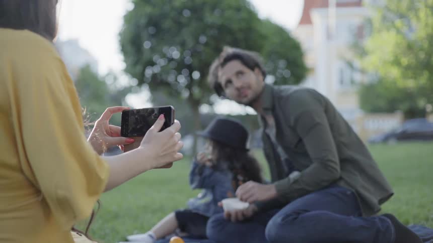A mother taking a photo of her husband and child during park picnic.
