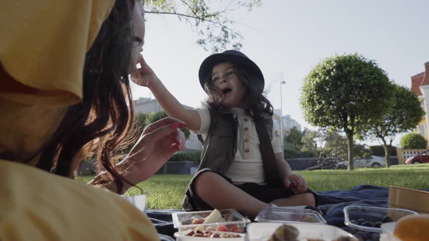 A boy is feeding his mother a bite of food as they picnic outside on a sunny day