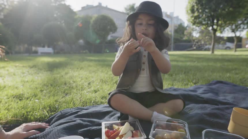 A young boy wearing a hat sitting on blanket eating snacks outdoors.