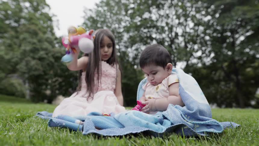 An older sister shaking a toy to play with baby on blanket.
