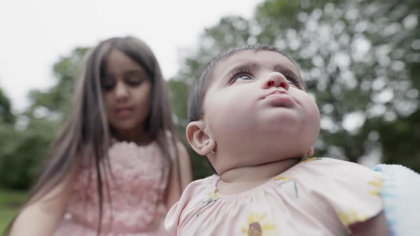 Adorable baby looking up curious while sitting near sister on picnic blanket.
