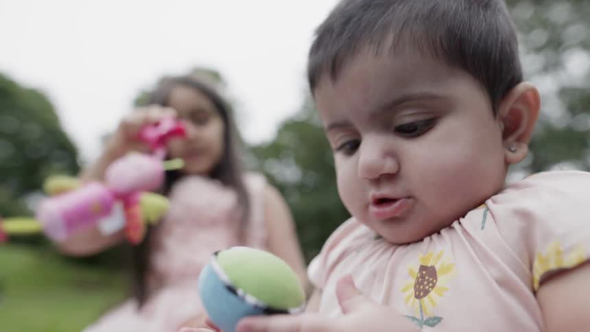A baby looking at a colorful toy while sister plays in background.