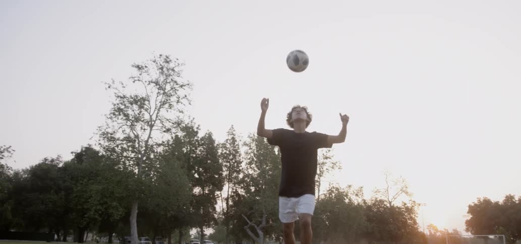Boy bounces soccer ball off his head in field at sunset