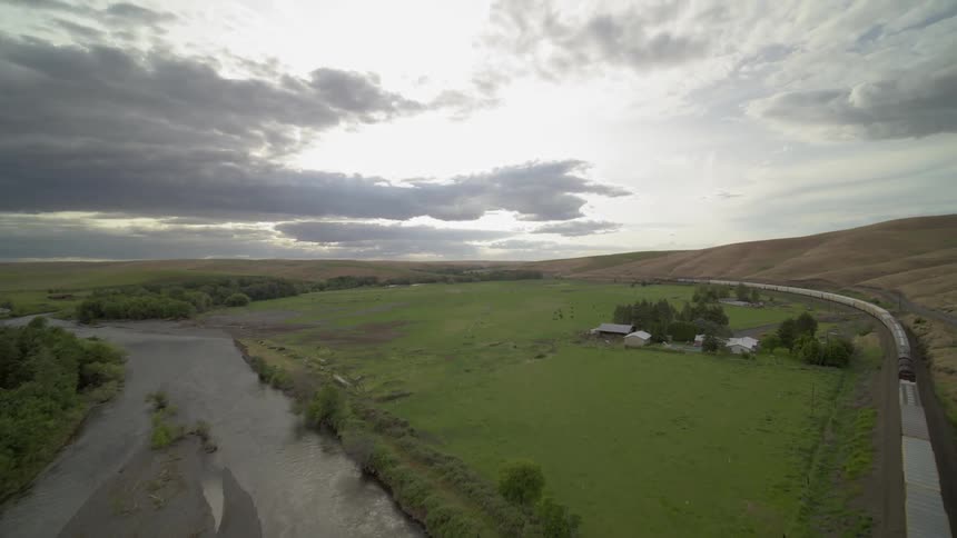 A freight train travels over a river next to farmland and grazing cattle