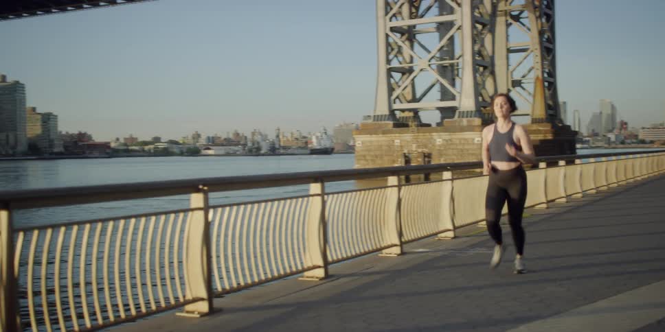 A woman jogs along a city harbor and past a bridge