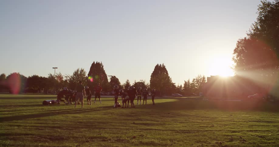 Football players practice on a grassy field at sunset