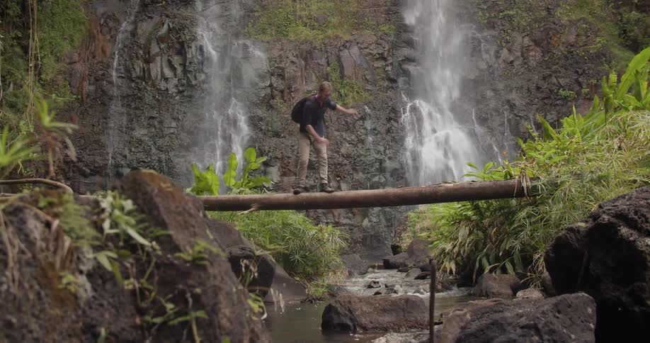 A man is nearly falling off a log suspended over a stream in a natural setting in daylight