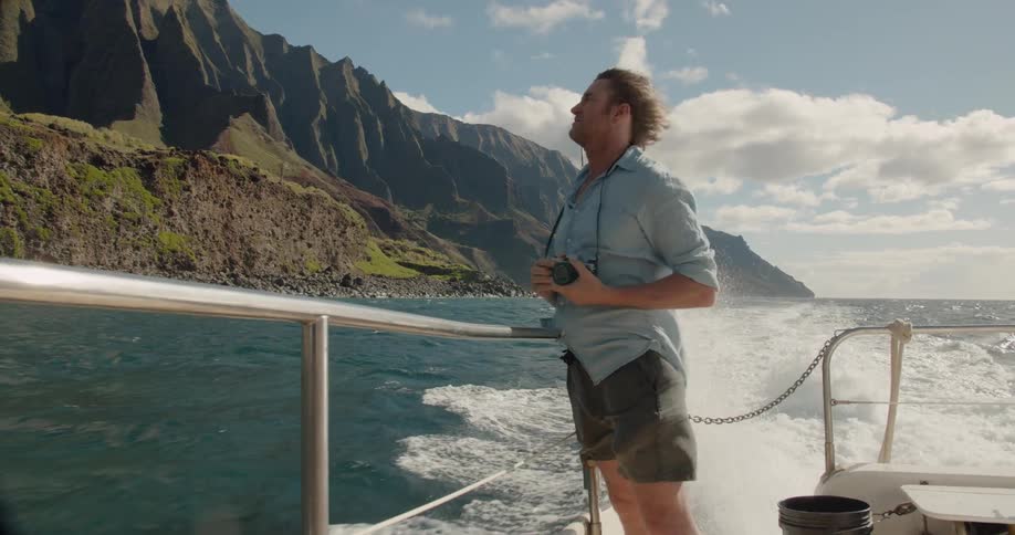 Man leans on the railing of a boat taking pictures of the shore