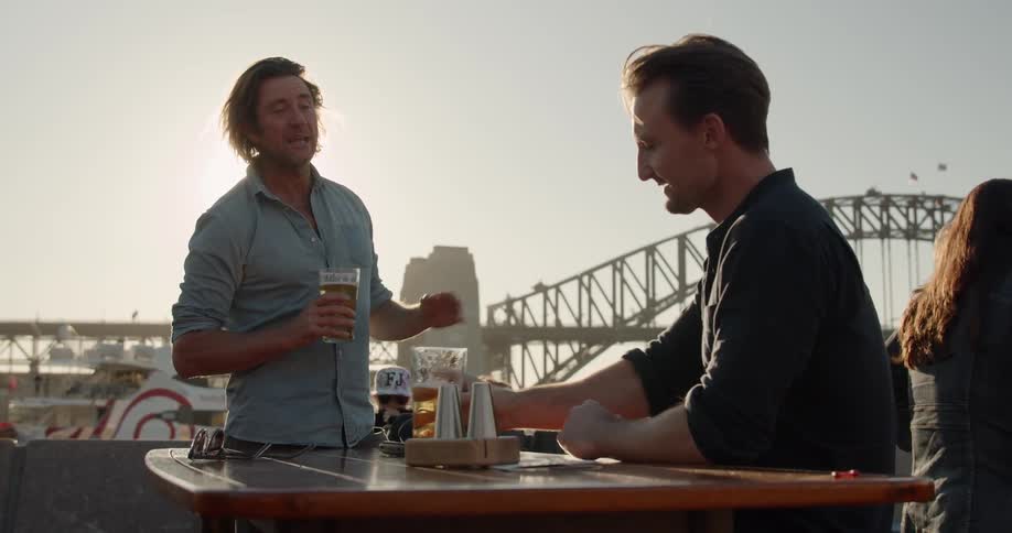 Two men share a beer outside near a bridge