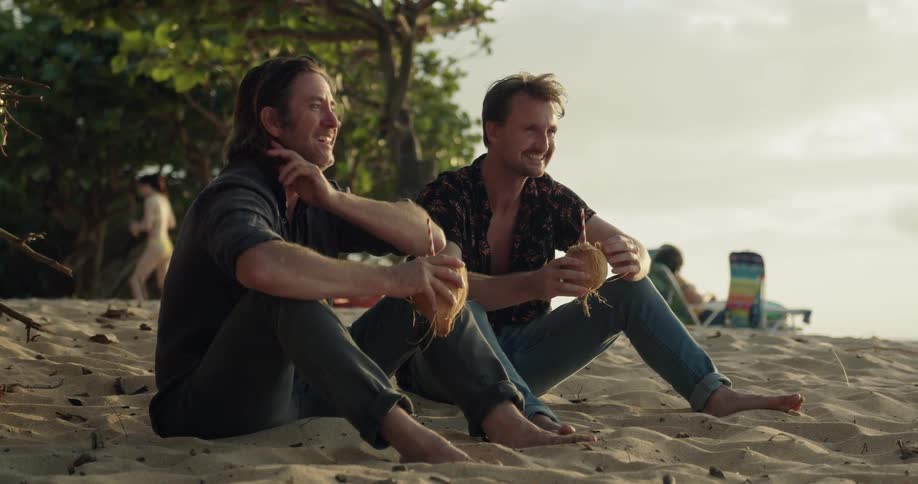 Two men drink from coconuts on the beach