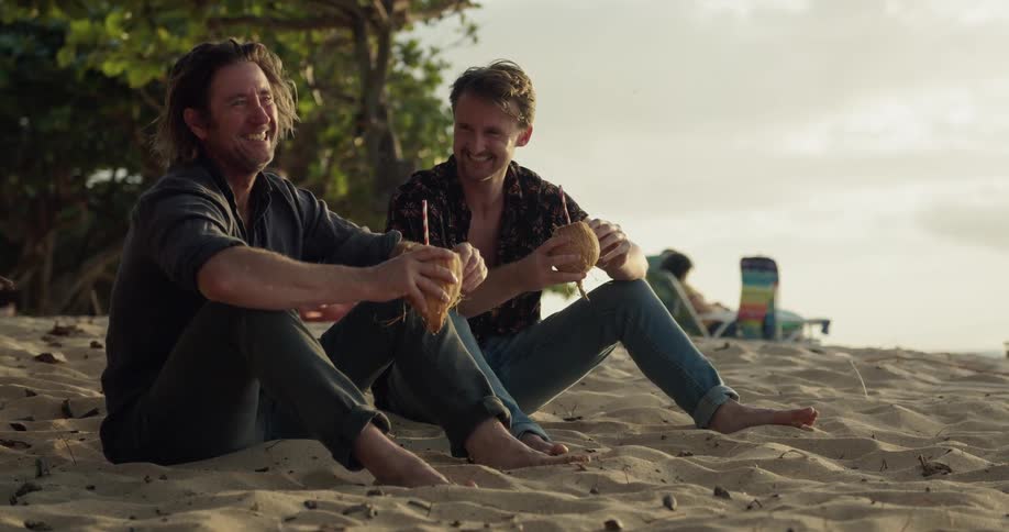 Two men drink from coconuts on the beach