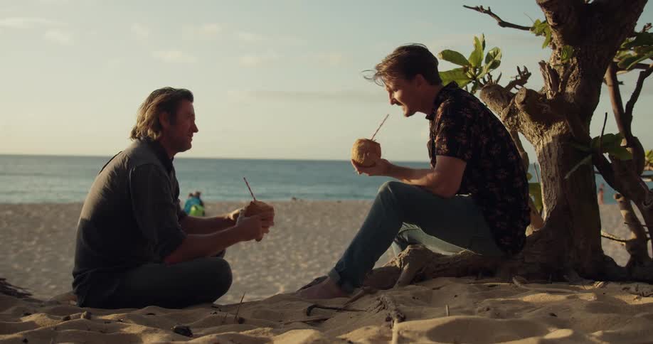Two men sit with coconut drinks on the beach