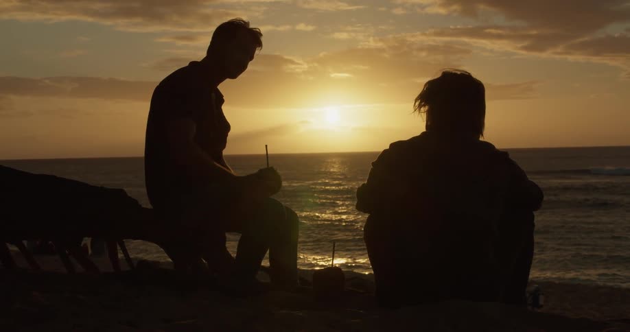 Two men talk on a beach at sunset