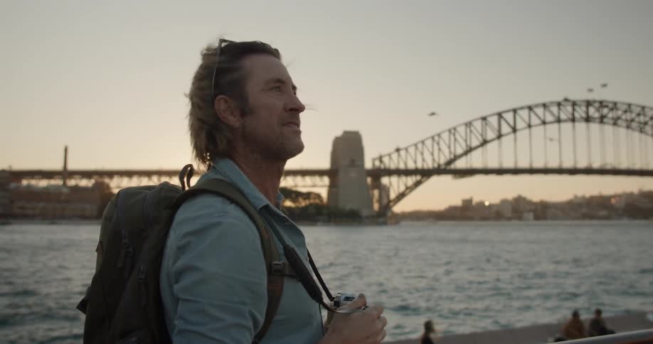A man walks near the water taking photos of a bridge