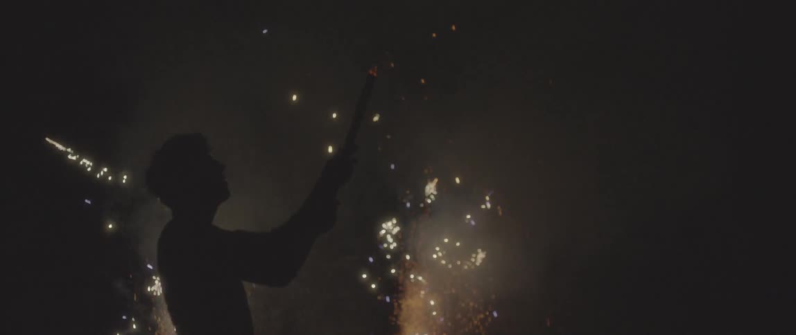 A man holding a roman candle while fireworks explode in the background