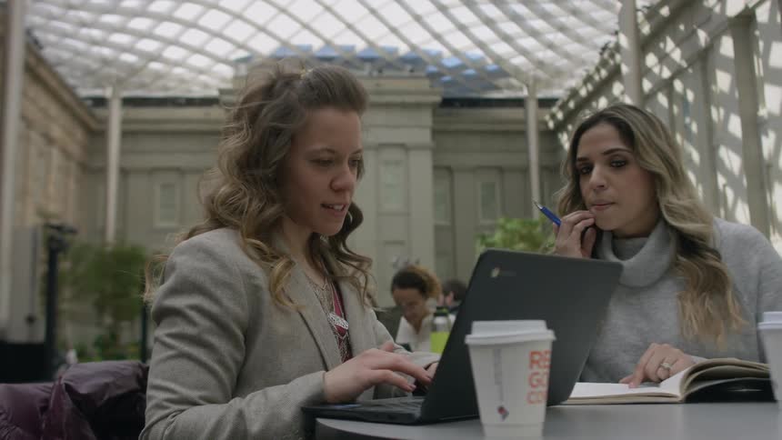 Two women sit in a courtyard with a laptop