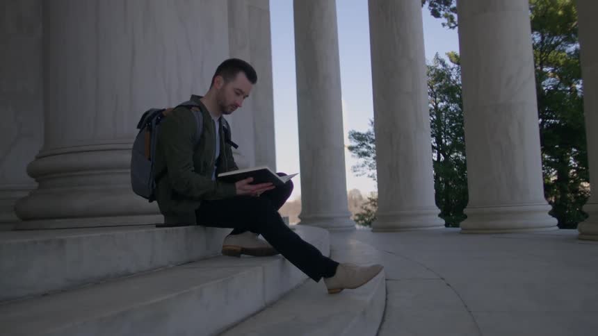 A man sits on the steps of a building with columns and reads a book