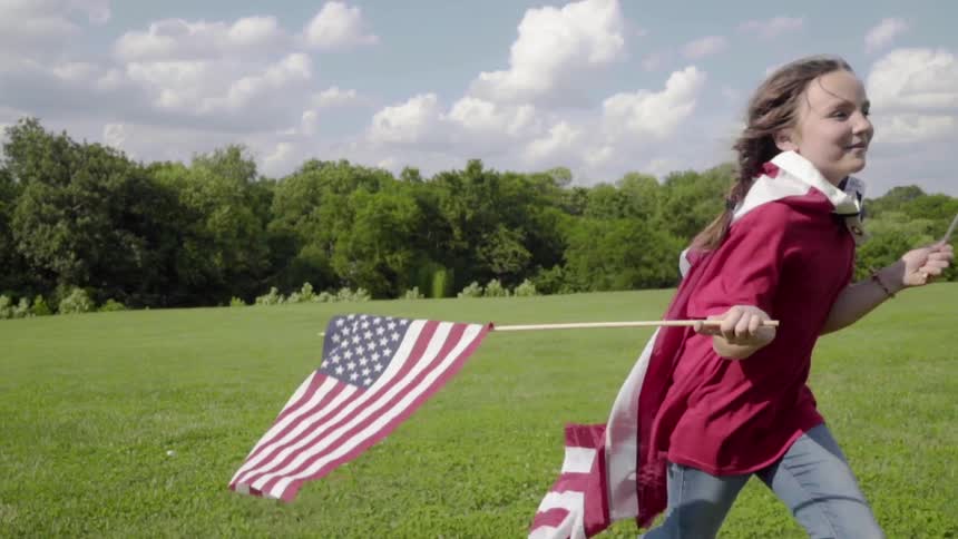 A girl wearing a flag as a cape runs through a field waving two small flags