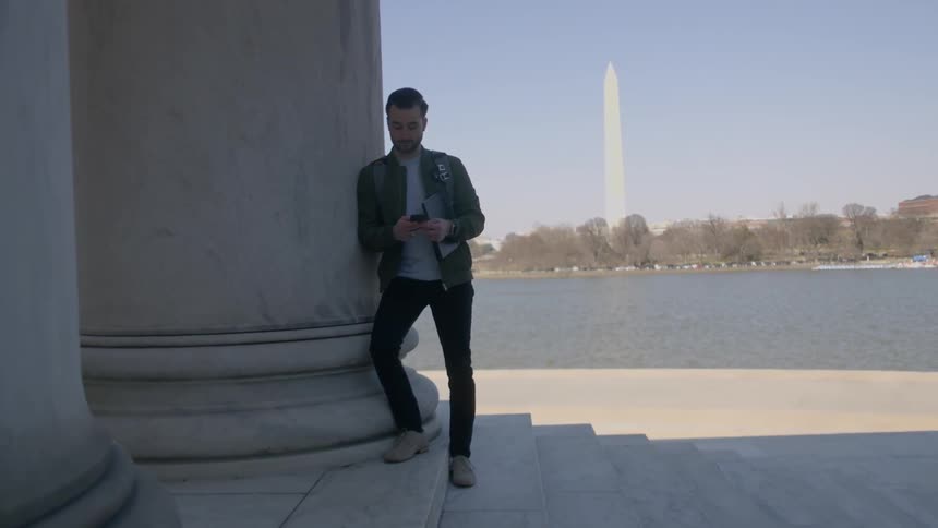 Man leans against a large pillar looking at his phone