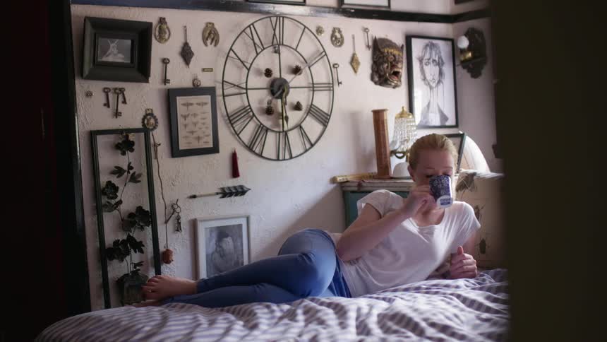 A woman sits in bed having a drink from a mug