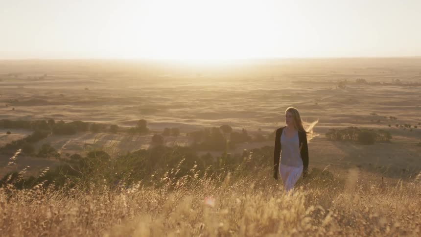 Woman is walking in weeds up hill at sunset on windy day