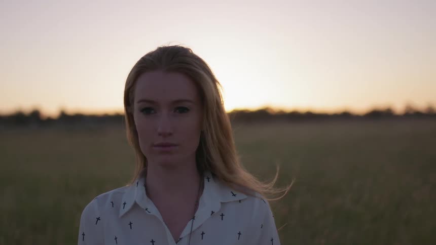 Woman is standing in field and staring off at sunset