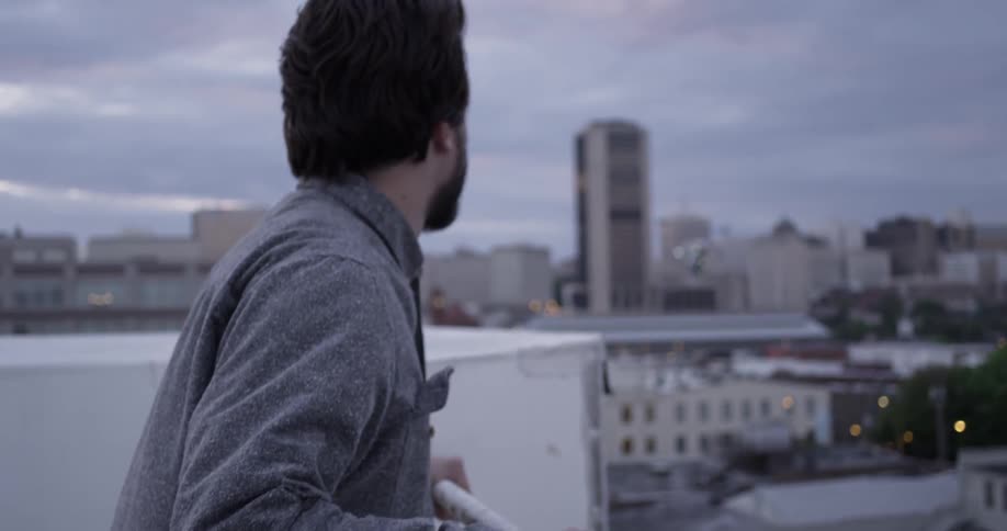 A bearded, brunette man walking up to a railing and grabbing it while he overlooks an urban area
