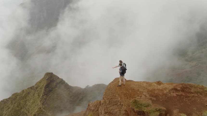 Two men stand on a ledge looking down at cloud covered peaks