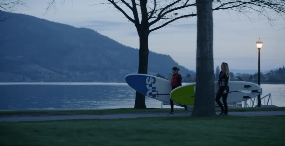 Two women walk on a path carrying paddle boards and oars