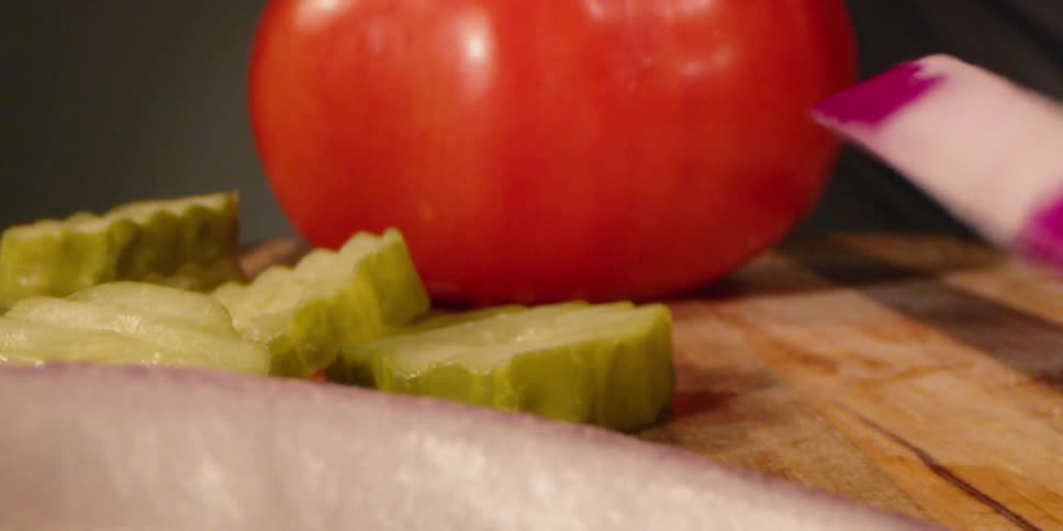 Sliced onion rings are tumbling onto a cutting board next to pickles and a tomato